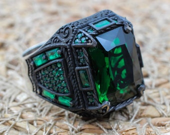 May include: A silver ring featuring a large, emerald-cut green gemstone. The ring has a dark, ornate setting with smaller green stones and decorative detailing. The ring is set against a textured, woven background.