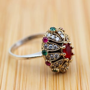 May include: A silver ring with a gold-toned setting featuring a large red gemstone surrounded by smaller white and green gemstones.