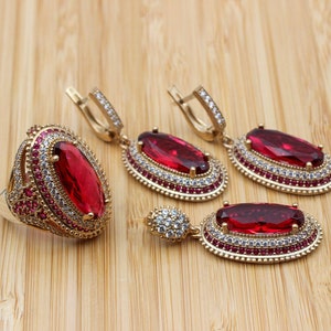 Peut inclure: Un ensemble de bijoux en pierres précieuses rouges comprenant une bague et une paire de boucles d'oreilles. La bague et les boucles d'oreilles sont dorées et présentent un design délicat avec de petites pierres blanches entourant les pierres précieuses rouges.