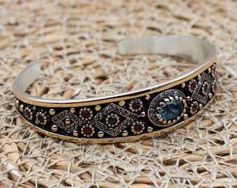 May include: Silver-tone cuff bracelet with a gold-tone and black band. The band is adorned with small red, green, and blue gemstones, and diamond-shaped patterns. The bracelet is set against a woven, tan background.