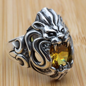 May include: A silver ring with a detailed lion head design. The lion's mouth is open, revealing a yellow gemstone.