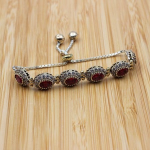 May include: Silver-toned bracelet with oval, red gemstone accents. The bracelet features a delicate chain and a sliding clasp for adjustable sizing. The gemstones are set in a decorative frame, adding to the bracelet's elegance.