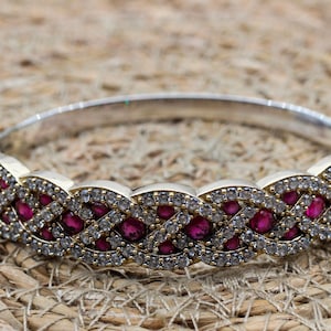May include: Silver-toned bracelet with a woven design. The bracelet features alternating sections of small, clear stones and larger, oval-shaped, red stones. The bracelet is set against a textured, tan background.