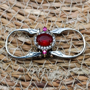 以下が含まれることがあります： 赤い宝石が小さな透明な宝石で囲まれた銀の指輪。指輪のバンドには装飾的なデザインが施されています。