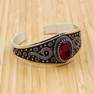 May include: A silver and gold cuff bracelet with an oval, red gemstone centre. The bracelet features intricate black and gold filigree detailing with small red and green accents. The bracelet is displayed on a wooden surface.