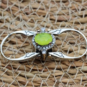 Può includere: Un anello argentato con una gemma centrale rotonda e verde chiaro, circondata da piccole pietre trasparenti. L'anello ha un design unico con bande curve che si estendono dal centro, su uno sfondo intrecciato.