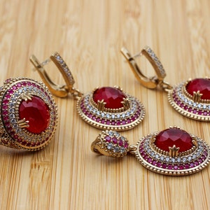 May include: A set of red gemstone jewellery featuring a ring, a pendant, and a pair of earrings. Each piece is adorned with a large red gemstone surrounded by smaller white and red gemstones.