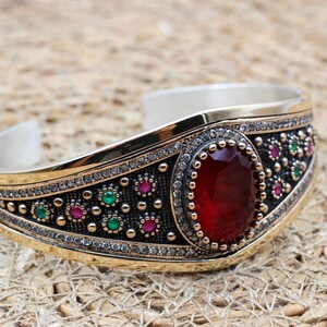 May include: A silver and gold cuff bracelet with a large oval red gemstone at the centre. The bracelet features a detailed design with small green, red, and clear stones. The bracelet is set against a textured, neutral background.