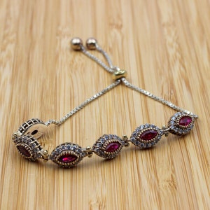 May include: Silver bracelet featuring oval ruby-red gemstones, each encircled by smaller clear stones. The bracelet has an adjustable chain. The jewellery is displayed on a light brown wooden surface.