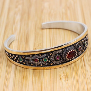May include: A silver bangle bracelet with a gold-toned band and intricate design featuring red, green, and white gemstones.