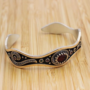 Puede incluir: Brazalete de puño plateado con un diseño ondulado. El brazalete presenta una piedra preciosa roja ovalada central, rodeada de piedras rojas, verdes y transparentes más pequeñas. Los detalles en negro y dorado añaden detalles.