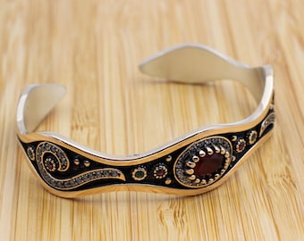 Könnte beinhalten: Silberfarbenes Armreif-Armband mit gewelltem Design. Das Armband hat einen zentralen ovalen roten Edelstein, umgeben von kleineren roten, grünen und klaren Steinen. Schwarze und goldene Akzente ergänzen das Design.