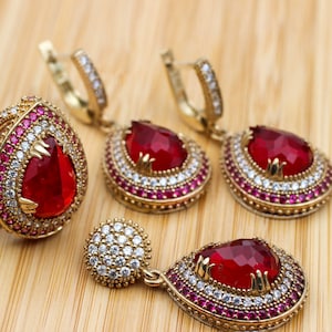 May include: A gold-coloured jewellery set featuring a ring and earrings with a teardrop-shaped, faceted red gemstone. The ring and earrings are adorned with smaller white and red gemstones, set against a light wooden surface.