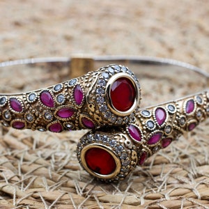 Könnte beinhalten: Silberfarbenes Armband mit aufwendigen goldfarbenen Details. Das Armband hat mehrere rote und klare Edelsteine. Das Design umfasst einen großen, runden roten Edelstein an jedem Ende.