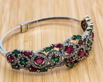 Pode incluir: Pulseira de prata com uma peça central decorativa com pedras preciosas vermelhas e verdes, acentuada com pequenas pedras transparentes. A pulseira está sobre um fundo de grão de madeira clara.