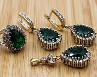 Könnte beinhalten: Ein Schmuckset mit einem Ring, Ohrringen und einem Anhänger. Die Teile haben eine goldfarbene Metalleinfassung mit einem tropfenförmigen, dunkelgrünen Edelstein, der von kleinen, klaren Steinen umgeben ist. Der Schmuck wird auf einer hellbraunen Holzoberfläche präsentiert.