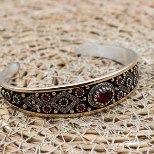 Può includere: Bracciale a polsino in argento con un design ornato. Il bracciale presenta una fascia dorata con accenti neri, adornata con pietre preziose rosse e verdi e motivi a forma di diamante. Una grande gemma rossa ovale è centrata.