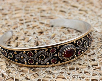 Könnte beinhalten: Silbernes Armreif-Armband mit einem verzierten Design. Das Armband hat ein goldfarbenes Band mit schwarzen Akzenten, verziert mit roten und grünen Edelsteinen und rautenförmigen Mustern. Ein großer, ovaler roter Edelstein ist mittig angeordnet.