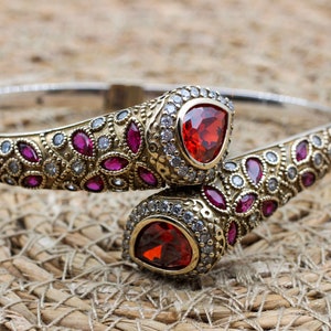 May include: A silver and gold-toned bracelet with a hinged design. The bracelet features a detailed pattern of small, round and oval-shaped gemstones in red and clear colors. Two large, triangular red gemstones are set at the ends.
