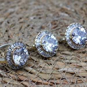 May include: Three gold rings with large clear cubic zirconia stones set in a halo of smaller stones. The rings are on a woven brown background.