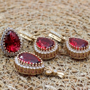 May include: A jewelry set featuring a ring and earrings with teardrop-shaped, deep red gemstones. The ring and earrings are accented with small, clear stones and gold-colored metal. The jewelry is displayed on a woven, tan surface.