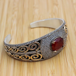May include: Silver cuff bracelet with a large, oval, red gemstone. The bracelet has detailed embellishments with small, clear stones and gold-coloured scrollwork. The open design allows for easy wear, with a circumference of approximately 17 cm.
