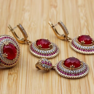 May include: A gold-toned jewelry set featuring a ring and earrings, each adorned with a large, oval, red gemstone. The ring and earrings are embellished with smaller red and white gemstones, creating a luxurious look. The set is displayed on a wooden surface.