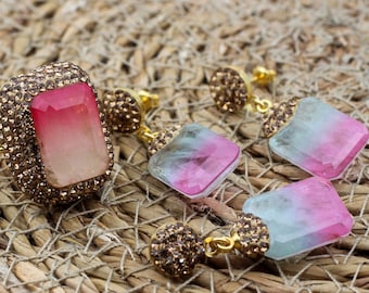 Puede incluir: Un conjunto de anillo y pendientes dorados. El anillo presenta una gran piedra rectangular rosa y blanca rodeada de pequeños adornos brillantes. Los pendientes tienen piedras rosas y azules similares, con adornos a juego.