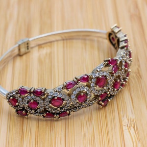 May include: Silver hinged bracelet with a decorative band. The band features oval and round red gemstones, surrounded by smaller clear stones. The bracelet is set against a light brown wooden surface.