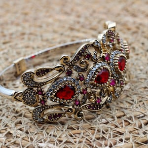 May include: Ornate silver and gold-toned bracelet with a detailed design. The bracelet features large, oval-shaped red gemstones surrounded by smaller, clear and red accent stones. The intricate metalwork has a filigree style, creating a luxurious appearance.