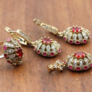 Peut inclure: Un ensemble de bijoux dorés avec des pierres rouges et blanches. L'ensemble comprend une bague, une paire de boucles d'oreilles et un pendentif.