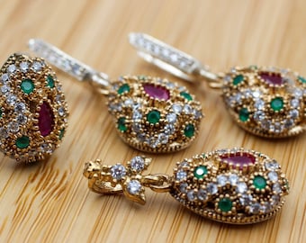 Könnte beinhalten: Ein goldfarbenes Schmuckset mit einem Ring und Ohrringen. Ring und Ohrringe haben ein tropfenförmiges Design, verziert mit roten, grünen und weißen Edelsteinen. Der Schmuck liegt auf einer hellbraunen Holzoberfläche.