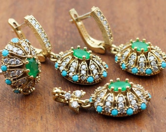 Könnte beinhalten: Ein goldfarbenes Schmuckset mit einem Ring und Ohrringen. Der Ring und die Ohrringe haben einen zentralen grünen Stein, umgeben von weißen und türkisfarbenen Steinen. Der Schmuck liegt auf einer Holzoberfläche.