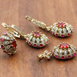 Puede incluir: Un conjunto de joyas doradas con un anillo y dos pares de pendientes. Cada pieza tiene una piedra preciosa roja central rodeada de piedras blancas y rojas más pequeñas. Las joyas se exhiben sobre una superficie de madera.