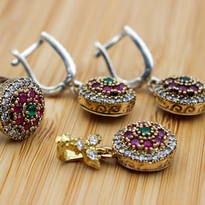 May include: A silver ring and two pairs of earrings with a gold and silver design. The jewelry features a central cluster of red and green gemstones, surrounded by smaller white stones. The pieces rest on a light brown wooden surface.
