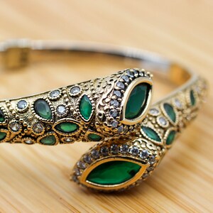 May include: A gold-coloured bracelet with a snake design. The bracelet features emerald-green and clear gemstones. The snake's head and tail are adorned with large, oval-shaped green stones. The bracelet is set on a wooden surface.