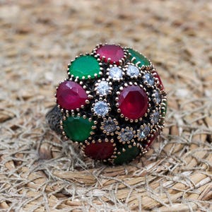 May include: A dome-shaped ring featuring a cluster of round-cut red and green stones interspersed with small, clear white stones. The setting is a dark, oxidized metal with a textured, intricate pattern visible on the band and the base of the setting.