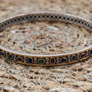 May include: A hinged bangle bracelet featuring a metallic band with a central black inlay. The inlay is decorated with a repeating pattern of oval-shaped red, blue, and green stones, interspersed with small clear stones set in a gold-tone frame.
