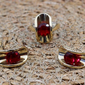 May include: A jewelry set consisting of a ring and two matching earrings. Each piece features a polished gold-tone metal frame with a curved, open-ended design that cradles a single, round, faceted deep red gemstone in the center.