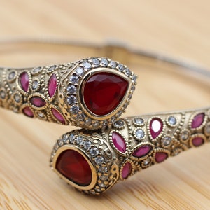 May include: A silver-toned cuff bracelet with ornate gold-colored detailing. The bracelet features two large, teardrop-shaped, deep red gemstones, surrounded by smaller red and clear stones. The design is symmetrical and elegant.