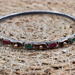 May include: A circular bangle bracelet featuring a dark oxidized silver band with alternating oval-shaped green and red gemstones. The metalwork includes decorative gold-toned accents and intricate filigree patterns between each stone setting.