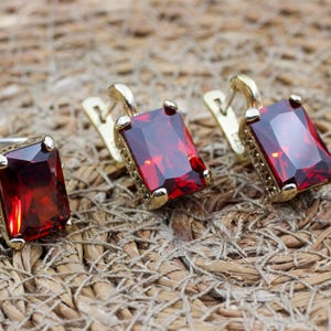 May include: A jewelry set consisting of a ring and two matching earrings, each featuring a large, rectangular, deep red faceted gemstone set in a gold-toned metal frame with intricate side detailing on the ring and earring settings.
