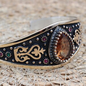 May include: A cuff bracelet featuring a central pear-shaped amber-coloured stone surrounded by small clear crystals. The metal band has a dark oxidised finish with gold-tone accents, intricate scrollwork, and small embedded red and green stones.