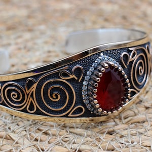 Puede incluir: Un brazalete tipo brazalete de plata con una piedra preciosa roja en forma de pera rodeada de pequeñas piedras transparentes. La superficie metálica tiene un acabado oxidado oscuro con patrones de espirales y bordes dorados pulidos.
