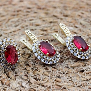Puede incluir: Un conjunto de joyería compuesto por un anillo y dos pendientes a juego, cada uno con una gran piedra roja de corte ovalado rodeada por un doble halo de pequeñas piedras transparentes, en metal dorado con un patrón de greca en los aros.