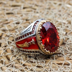 May include: A silver and gold ring with a large, oval, red gemstone. The ring has a red enamel band with a gold sword design. The ring is set on a woven, tan surface.