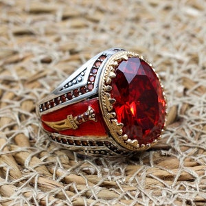 May include: A silver ring featuring a large red gemstone and an intricate design of a sword on the band.