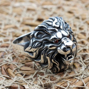 Può includere: Un anello con leone in argento con dettagli intricati. La testa del leone presenta una criniera dettagliata, bocca aperta e occhi di pietre preziose rosse. L'anello è posto su uno sfondo intrecciato e strutturato.