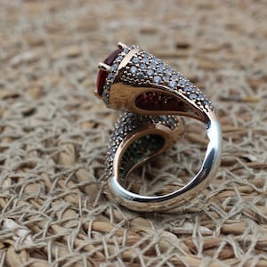 May include: A silver ring featuring a large red gemstone and smaller clear gemstones. The ring has a twisted band design.