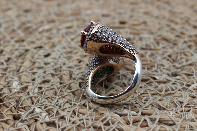 May include: A silver ring featuring a large red gemstone and smaller clear gemstones. The ring has a twisted band design.